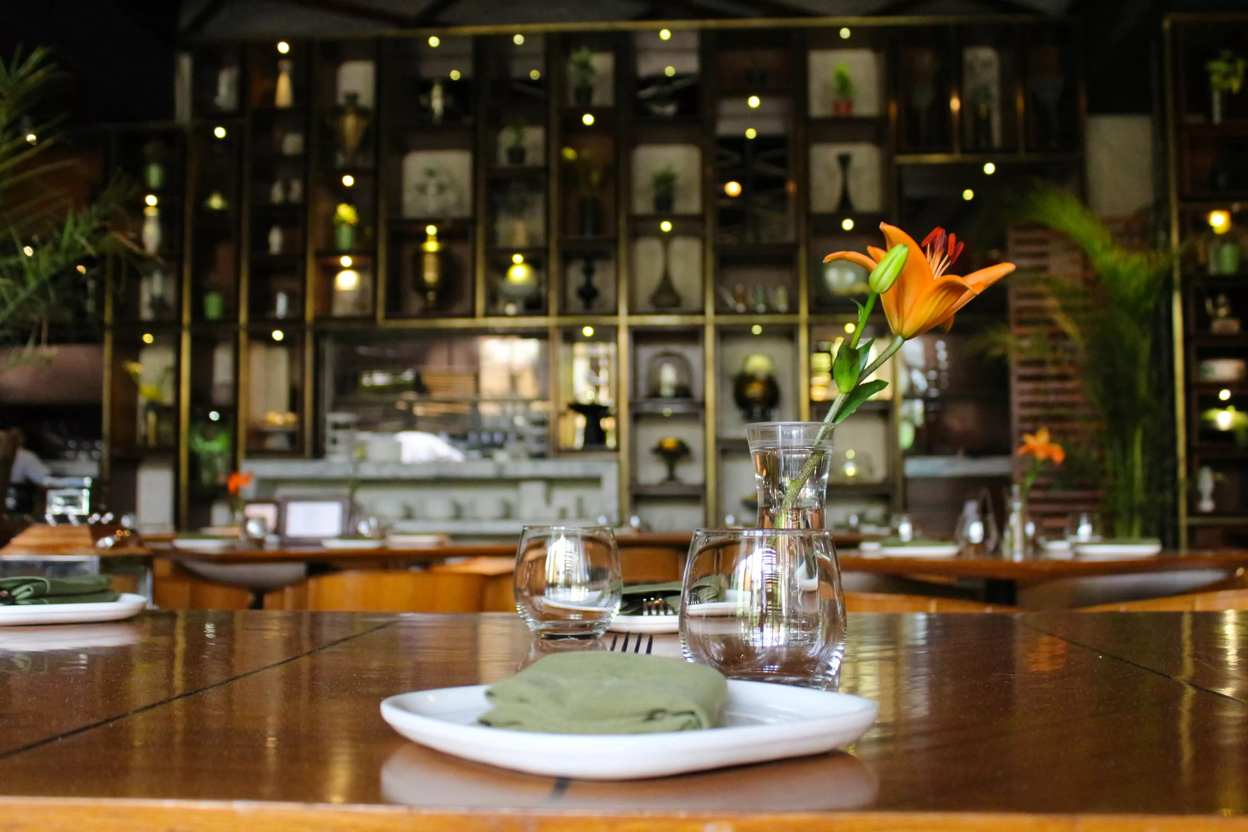 A restaurant table with glasses, plate, orange Lilly, and bar with drinks